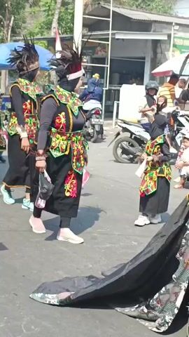 The video shows a procession of people, primarily women and children, dressed in elaborate traditional costumes. They are walking along an asphalt road, likely participating in a parade or cultural event. The costumes are predominantly black with colorful, ornate detailing on the vests and headpieces, which are adorned with feathers. Some individuals are wearing hijabs underneath their headwear. Several people are holding small flags, some of which appear to be Indonesian flags. In the background, there are buildings, tents, a motorcycle, and other onlookers, suggesting an outdoor public gathering. The atmosphere seems festive and organized.