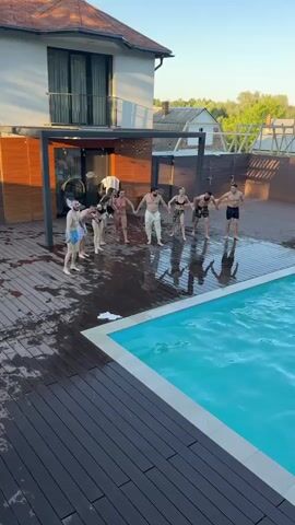 A group of approximately ten young adults, dressed in swimwear, stand in a line on a wet wooden deck next to a swimming pool. They are holding hands and appear to be running towards the pool. One person jumps into the pool, creating a large splash. The scene takes place outdoors during daylight, with a modern house and trees visible in the background.
