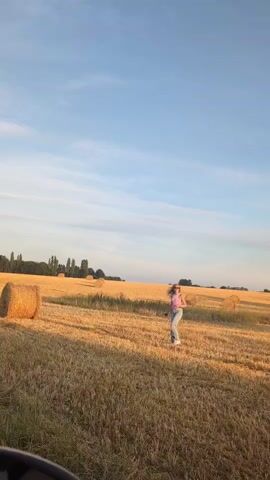 The video shows a person standing in a field of dry grass with several hay bales visible in the background. The sky is clear and blue with some clouds. The person is wearing a pink shirt and blue jeans and appears to be posing for a photo. The overall impression is one of a rural landscape during daytime.