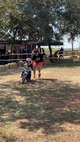 A child wearing a helmet and protective gear is riding a small blue dirt bike on a grassy field. An adult man, wearing a black t-shirt and pink shorts, is walking alongside the child, holding the bike for support.  Hay bales and white tape mark the perimeter of a makeshift track. Behind the track are trees, tents, and several other people. The ground is covered with grass and dry patches of dirt. The overall setting appears to be an outdoor event or competition for children learning to ride dirt bikes.