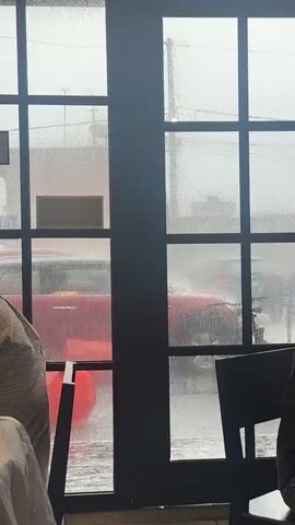 The video shows a view through a window with raindrops on it. Outside, a red car is parked on a street that appears to be flooded. Water is splashing up on the car, suggesting heavy rain and possibly wind. In the foreground, partially visible through the window, are the backs of two people sitting in chairs. One person is wearing a light-colored shirt, and the other is wearing a dark shirt. Through the window, a red sign with white lettering is visible, possibly indicating a business. The overall impression is of a stormy day.