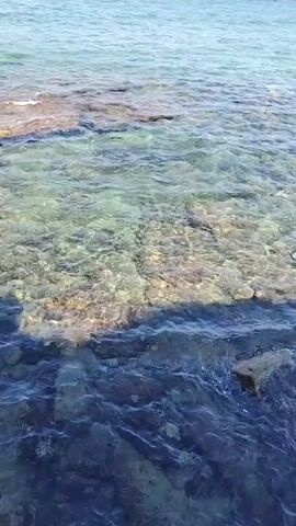 A video shows a body of water, likely the ocean, with a rocky shore. The water is clear, allowing the bottom to be seen in shallower areas. The water depth increases towards the right side of the frame, becoming darker blue. Sunlight reflects off the water's surface, creating shimmering patterns. The sky is visible in the distance, appearing clear and blue.