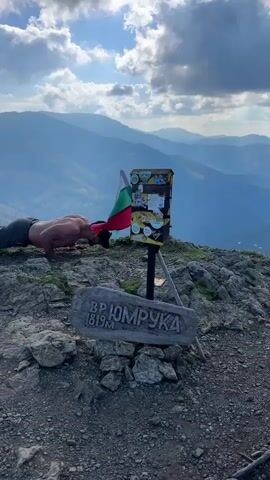 A man is doing push-ups on a rocky mountain peak, with a Bulgarian flag and a signpost behind him. The signpost has stickers and appears to be weathered. The man is shirtless and wearing shorts and a hat. The background shows rolling mountains under a cloudy sky.