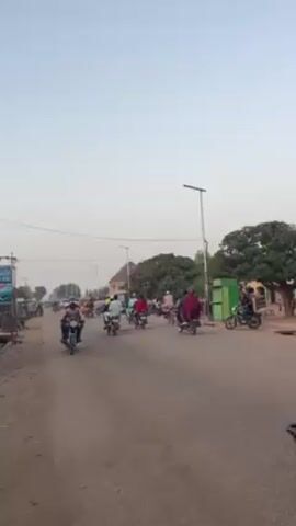 The video shows a street in what appears to be a town or city in Africa, with many people riding motorcycles. The road is paved but looks dusty, and there are buildings and trees along the roadside. The sky is overcast, suggesting either early morning or late afternoon. People are dressed in various clothing, some wearing traditional attire. There's a mix of motorcycles, and a red three-wheeled vehicle can also be seen. The overall impression is of a busy and active street scene in a developing region.