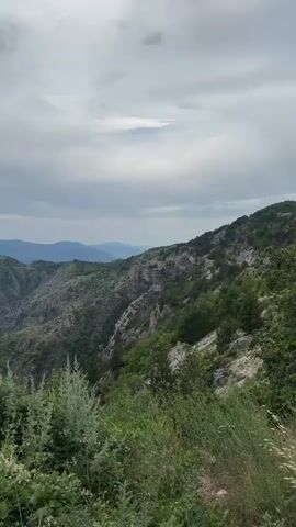 This is a video recording of a scenic mountain landscape in Kućišta, Montenegro. The camera pans across a vast expanse of rugged mountains covered in lush green vegetation. In the foreground, wild grasses and bushes are visible, some with small white flowers. 

The terrain is characterized by steep slopes, deep valleys, and dramatic rock formations, including sheer cliffs and prominent rock spires. The mountains in the distance appear hazy and are layered in various shades of green and blue, suggesting a significant depth to the landscape. The sky is overcast with a mix of gray clouds and some brighter patches, indicating a cloudy day, possibly with the sun attempting to break through. There are no people or man-made objects visible in the footage. The overall impression is one of natural beauty, wilderness, and a sense of awe inspired by the scale and grandeur of the mountains. The time of day appears to be daytime, given the ambient light. There is no visible text in the media file.