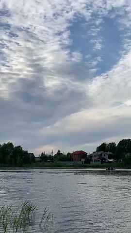 The video shows a body of water with some reeds in the foreground. In the background, there are houses with trees and a cloudy sky. A small boat with two people in it can be seen on the water. The lighting suggests it is either early morning or late evening.