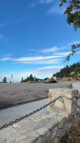 A truck with a trailer is parked in a paved area. A flock of birds is flying in the blue sky. There are trees and signs in the background. A stone wall with a chain is in the foreground.