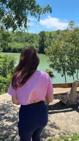 The video shows a woman standing on a stone surface overlooking a lake on a bright, sunny day. She is wearing a short pink t-shirt and dark blue leggings. She turns briefly towards the camera, revealing she's also wearing sunglasses. The lake is surrounded by green trees and hills, and there is a small, house-shaped structure floating on the water. A stone bench with an unusual design sits in the foreground. The camera pans slightly to the right, showcasing the view.