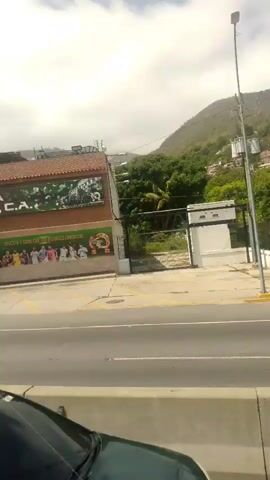 This video, filmed in Macuto, Venezuela, captures a view from a moving vehicle on a cloudy day. The scene is dominated by a multi-lane road with a concrete barrier separating the lanes. In the foreground, the roof of a dark green car is visible.

The mid-ground showcases a white sedan driving on the road. Beyond the road, a hillside is covered with lush green trees and scattered buildings, suggesting a residential area on the slopes. A metal fence runs along the edge of the road, separating it from the terrain beyond. A prominent lamppost stands near the road, and a utility pole with wires is also visible.

In the distance, hazy mountains form the backdrop to the scene. The overall atmosphere is tranquil, with no discernible emotions expressed. The weather appears overcast, with the sky filled with clouds. No text is clearly visible within the frames of this short clip.