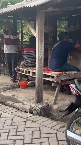 The video captures an outdoor scene under a rustic shelter with a red and white corrugated roof. In the foreground, a red scooter is parked on a dirt ground. A person wearing a blue long-sleeved shirt and jeans is squatting on a wooden bench to the right, seemingly holding a cigarette. Behind them, more foliage and trees are visible. The camera pans slightly to the left, showing the front of a black and white motorcycle. To the left of the frame, a man in a striped shirt and dark pants is walking away from the camera, gesturing with his left hand. Two other individuals are partially visible in the background, seated under the shelter. The ground is paved with interlocking stones in the lower portion of the screen, transitioning to dirt where the vehicles are parked. The overall environment appears to be a casual, outdoor gathering or rest stop.