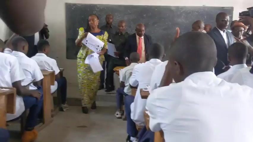 The video shows a scene in what appears to be a classroom. A woman in a colorful, long dress is standing and distributing papers to students sitting at desks. Several men in suits and other professional attire are standing behind the woman, observing. Some of the men are holding cameras. The students are wearing white shirts and are raising their hands, presumably to receive the papers. A chalkboard is visible in the background. The classroom appears to be simple, with wooden desks and a light-colored wall. The room is filled with people and activity, possibly indicating a formal visit or special event in the classroom.