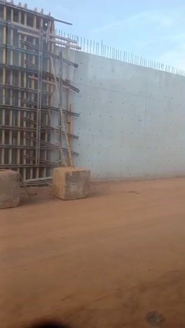 The video shows a construction site with a large concrete wall. Rebar protrudes from the top of the wall. Wooden scaffolding is attached to the left side of the wall. A concrete block sits at the base of the scaffolding. The ground is dirt. The sky is visible above the wall, with some clouds. A person is visible in the distance on the right side of the screen, walking.