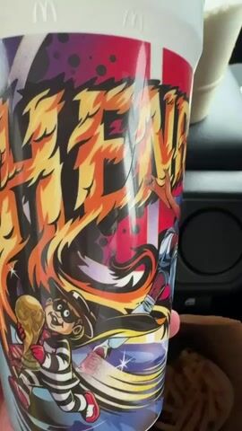 A person is holding a McDonald's cup with a colorful design. The design features illustrations of football players, a soccer ball, and the McDonald's logo. There are also graphics that appear to be flames and explosions. In the background, a brown paper bag with french fries and another McDonald's drink are visible. The cup is being rotated to show different parts of the design.