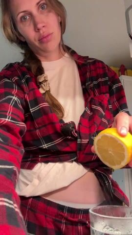 A woman is squeezing a half lemon into a glass of water with ice. The woman has light brown hair in a side braid and wears a red and black plaid pajama set over a white t-shirt and white underwear. She looks directly at the camera while squeezing the lemon. The glass of water with ice is placed on a surface in front of her. The background shows a white cabinet with some items on top.