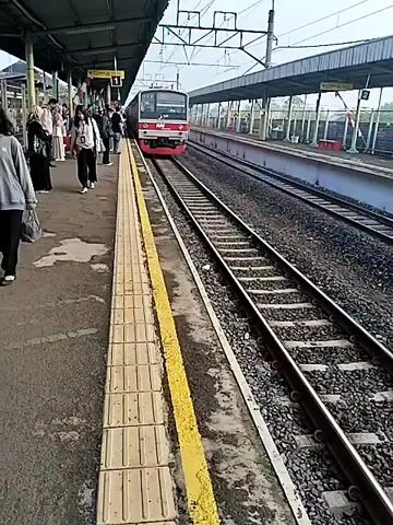 The video opens at a train station platform. A red train is arriving on the tracks to the right of the frame. Several people are standing on the platform to the left. The platform is concrete with yellow tactile paving along the edge. Above the platform is a covered waiting area with a metal roof. Overhead power lines are visible above the tracks. As the train continues to arrive, its side becomes visible, showing the word "SHARP" on one of the cars. People on the platform are looking at the arriving train. The camera pans slightly to follow the train as it moves past the platform.