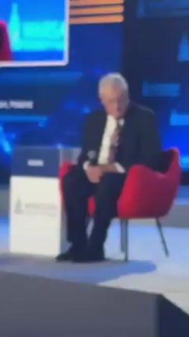 The video depicts an elderly man seated in a red armchair on a stage. He is wearing a dark suit with a white shirt and a red-striped tie. He holds a microphone in his right hand and looks down pensively, occasionally glancing to his left. The backdrop is a blue screen displaying the logo of the "Warsaw Security Forum," a conference held in Poland. A smaller screen in the background also displays the same logo. A white sign with the Kellogg logo and "Warsaw Security Forum" is positioned near the chair. The stage is covered with light blue fabric. The man appears to be either waiting to speak or reflecting on a question posed to him.
