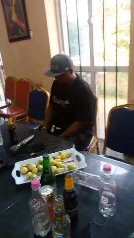 The video appears to show a social gathering or event.  

Several individuals are present.  One man wearing a hat and black shirt sits at a table laden with bottles (likely alcoholic beverages) and fruit.  Other people are standing and walking around the room.  One woman is wearing a white outfit, while another is wearing a black and yellow striped outfit. The room has several chairs around tables, a window with bars, and artwork on the wall.  Music is audible. The camera is unsteady and moves frequently.