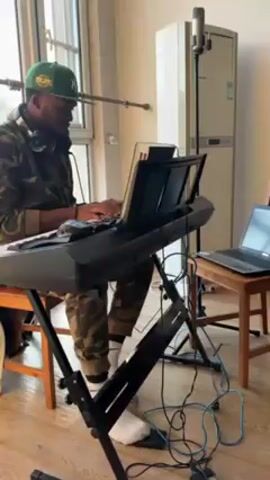 A man is playing a keyboard in a room. He is wearing a green baseball cap, a camouflage jacket, and white socks. Headphones are around his neck. The keyboard is on a stand, and a laptop is on a small wooden chair next to it. A microphone is on a stand behind the keyboard. There's an air conditioning unit along the wall. The floor is made of light-colored wood. A window is visible in the background. The room appears to be a home studio setup. Wires connect the keyboard to the laptop. The man is focused on playing the keyboard.