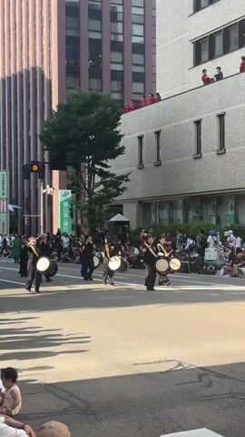 A daytime wide shot shows a Japanese street parade with approximately 10 performers in black clothing with yellow sashes, walking forward with taiko drums and flutes, in a straight line on a city street. Spectators sit on the curbs, and some people stand on a building's rooftop balcony on the right. Tall buildings line both sides of the street. Shadows of unseen objects are visible in the foreground.