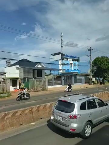 The video shows a street scene with buildings and vehicles. On the left, there is a house and a telecommunications tower. A person on a scooter is traveling on the road. In the center, there is a blue building with signs that appear to be for a telecommunications company, possibly "Vinaphone." There is also a large green tree. A silver SUV is driving on the road, and there are other buildings and structures along the street. The sky is partly cloudy.