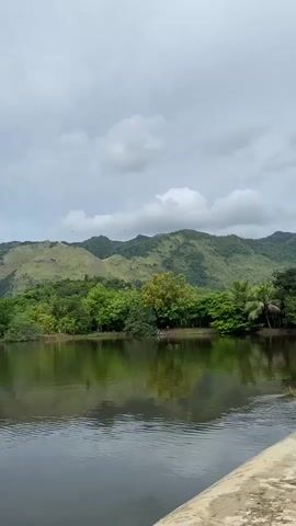 The video shows a serene landscape with a body of water in the foreground and lush green mountains in the background. The sky is overcast with scattered clouds. The water is calm, reflecting the trees and mountains. There are trees and foliage along the bank of the water. The overall impression is one of natural beauty and tranquility.
