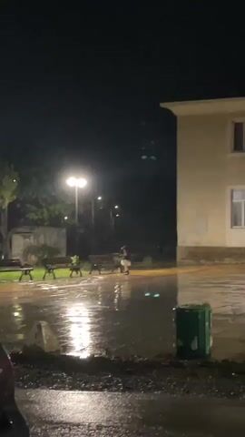 At night, a person sits on a bench in a rain-soaked square under the glow of a street light. The scene is illuminated by the light reflecting off the wet ground. There are several empty benches and a building in the background. A green trash can is visible near the foreground. A car is parked near the bottom left corner of the frame.