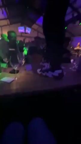 The video shows a person standing on a table at an event. The person is wearing black pants and black-and-white sneakers. The room is brightly lit with green and blue lights, giving a party atmosphere. There are other people sitting at tables around the room, watching the person on the table. There are glasses and bottles on the table. A band can be seen in the background.