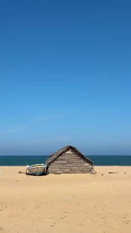 The video shows a small, thatched-roof hut situated on a sandy beach. Beside the hut is a white and blue boat. The ocean is visible in the background, meeting a clear blue sky. The scene is still and peaceful, with no people or discernible activity. The overall environment is a tranquil beach setting.