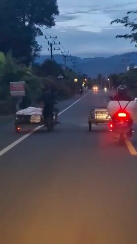 The video shows two motorbikes with attached sidecars driving on a two-lane asphalt road at dusk. The camera is seemingly mounted on a vehicle following the motorbikes. The first motorbike, seen on the left, carries several white bags, likely full of produce or goods, in its sidecar. The second motorbike, on the right, has a metal-caged sidecar, possibly transporting animals or crates. Both vehicles have their taillights on, casting red glows on the road. Overhead power lines and poles are visible along the road, which is flanked by trees and distant hills under a cloudy sky. The overall scene suggests a rural or semi-rural setting as the day transitions to night.