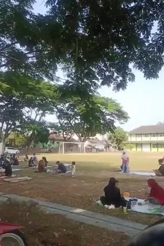 The video shows an outdoor scene on a sunny day, likely a park or school grounds, with a large tree providing shade in the foreground. Several groups of people are spread out on the grass, sitting on blankets or mats. Some individuals appear to be picnicking, with food and drinks visible. In the background, there is a sports field with goalposts and a building that resembles a school. The overall atmosphere seems relaxed and casual, with some adults and children present. The camera pans slowly, offering a panoramic view of the gathering.