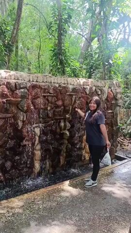 The video shows a woman standing near a small artificial waterfall. The waterfall is made of layered stones, with water flowing down its surface into a narrow trough at the base. The woman is wearing a dark-colored shirt, dark pants, and gray sneakers. She is smiling and waving at the camera. In her other hand, she is holding a white plastic bag. The area surrounding the waterfall is a park-like setting, with trees and green vegetation visible in the background. The ground is paved with gray tiles. The lighting suggests it is a bright day.