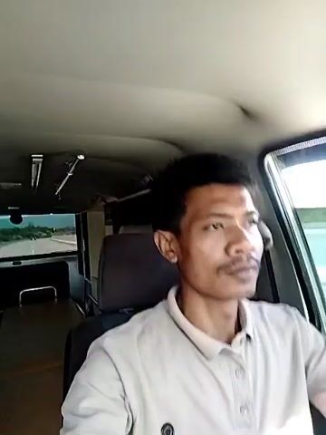 A man is driving a van on a road. The man is wearing a beige polo shirt. Inside the van, behind the driver’s seat, is a stretcher. The road can be seen through the back windows.