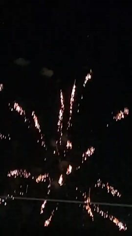 The video shows a fireworks display at night. Various fireworks are seen exploding in the dark sky, illuminating the scene with bright colors such as white, pink, and green. Some fireworks create a fan-like or spherical shape with trails of light, while others appear as streaks or bursts. In the foreground, a dark silhouette of what appears to be a building with windows is visible against the illuminated sky. The display appears to be taking place outdoors, possibly in a residential or recreational area. The overall atmosphere is festive and celebratory.