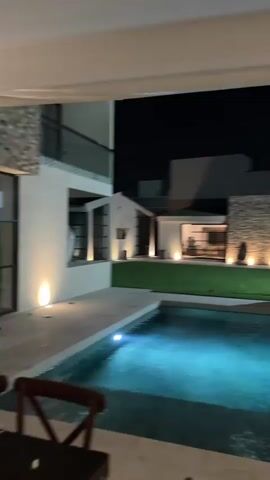 The video shows a luxurious backyard scene at night. There is a swimming pool illuminated with blue lights, surrounded by a tiled deck. Beyond the pool is a well-manicured lawn and a modern house with large windows and exterior lighting. Several people are standing on the lawn, some of them interacting. Music is playing in the background. The camera is positioned under a covered patio or porch, providing a partial view of the scene.