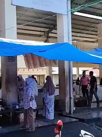 The video depicts a bustling outdoor market setting. Numerous individuals are visible, some standing and observing, others actively engaged in activities related to the stalls. Several stalls are present, with some displaying fish and others showcasing cuts of meat hanging from hooks.  Blue and orange tarps are strung up to provide shade for the vendors and customers. The market appears to be located near a building with white pillars and a corrugated metal roof. The ground is wet, suggesting recent cleaning or rain. One individual is seen smoking a cigarette. Overall, the video captures the lively atmosphere of a traditional market.