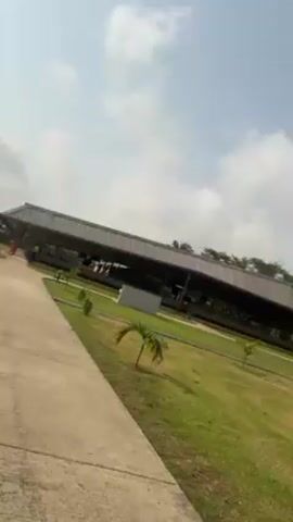 The video shows a sunny day at an outdoor location with well-maintained green grass, trees, and a long, covered walkway or building structure. The camera pans slightly to the right, revealing a concrete path leading toward the walkway. There is a concrete structure that looks like a hand washing station to the left of the path. In the distance, a person wearing a yellow shirt can be seen walking on the path towards the covered walkway. The sky is blue with scattered white clouds.