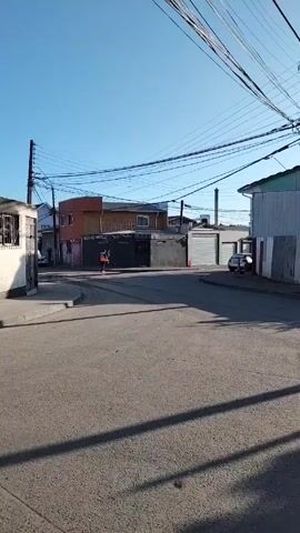 The video shows a street in what seems to be a small town, perhaps in a rural area. The sky is clear and blue. The road is paved and empty, save for the shadow of the videographer. Several buildings line the street. One structure displays visible fire damage, blackening the facade above the windows and around the door. A flag, red and white, is visible through one of the windows. Graffiti is scrawled on the building. The camera pans from left to right, capturing the street and the buildings. The video seems to be a brief recording of the location, possibly as a form of documentation or record-keeping.
