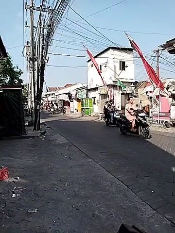 A street scene is captured under a clear blue sky with a dense network of overhead wires dominating the upper left quadrant of the frame. The street is paved with asphalt and appears to be a busy thoroughfare, lined with buildings on both sides. Red and white flags, likely national flags, are displayed prominently on poles along the right side of the street, suggesting a possible national holiday or celebration. Several people are seen on motorcycles, navigating the street. On the left side of the street, there is a dark structure, possibly a shop or stall, partially covered with black tarp, and some debris is scattered on the ground in the foreground. The overall atmosphere suggests a typical day in a bustling urban or semi-urban environment.