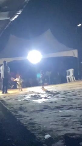 A group of people are gathered under a large white tent at night, illuminated by a bright light source. In the foreground, the ground appears to be dirt and possibly wet. Two children are in the center of the scene, one is crouched down while the other stands. A man in a suit is standing to the left of the children. Other figures are visible in the background, some seated and some standing. The overall impression is of an outdoor gathering or performance.