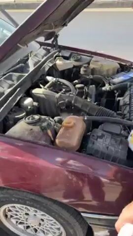 A man films the open hood of a burgundy car, from which steam emerges near the coolant reservoir. The camera zooms closer to the coolant reservoir before the camera pans back to show the man filming, who is standing next to another man. The second man is holding a bottle. The car is parked on the side of a paved road. The background includes a bright blue sky, desert hills, and passing traffic.