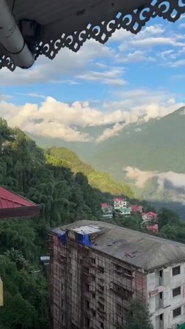 A video pans across a mountainous landscape. In the foreground, an unfinished concrete building with scaffolding and a blue tarp on its roof is visible. Below the building, a cluster of colorful houses dot a steep, tree-covered hillside. The background is dominated by lush green mountains, with some peaks obscured by white clouds. The sky is blue with scattered white clouds. The camera appears to be positioned on a balcony or elevated structure, as evidenced by decorative metalwork at the top of the frame.