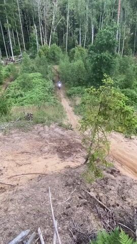 A person is riding a dirt bike up a sandy hill in a forest. The rider is wearing a blue jersey and a helmet. The hill is covered in sand and dirt, with some trees and vegetation growing on the sides. The forest is dense with tall trees. The video is taken from a high vantage point, looking down on the hill and the rider.