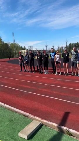 Here is a description of the video:
The video shows a group of people lined up on a red running track. The people appear to be preparing for a race or event. Some are wearing athletic clothing, while others are in casual attire. A person wearing a neon green shirt and weighted vest approaches the starting line. The background includes a clear blue sky, trees, and some buildings. The scene takes place outdoors on a sunny day.