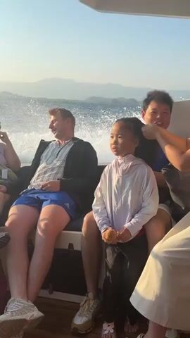 A group of people are on a boat moving through the water. A man in a blue shirt is sitting on the left side of the boat and is looking to the left. A young girl is sitting in the middle of the boat and is looking forward. A man in a blue shirt is behind the girl and is braiding her hair. The water is splashing up on the right side of the boat. The sun is shining and the sky is clear.
