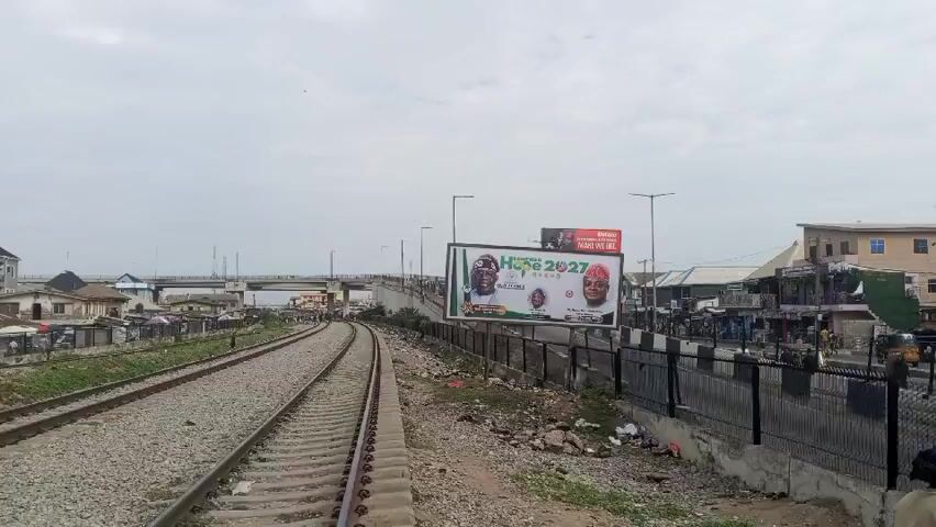 The video shows a view from train tracks in what appears to be a bustling urban area. The railway tracks curve slightly to the left, with gray gravel surrounding them. A billboard dominates the right side of the frame, featuring political figures and the text "HOPE 2027". Behind the billboard, there is a road with vehicles, including a large white truck, and pedestrians visible. Buildings and shops line the road. On the left side of the tracks, there are glimpses of houses and market stalls with people moving around. An overpass is visible in the distance. The sky is overcast and gray. A woman wearing a blue headwrap walks along the side of the road.