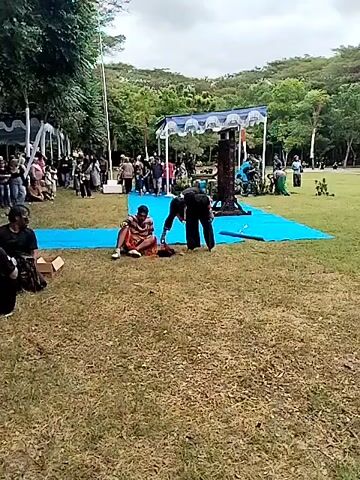 The video shows an outdoor gathering of people, likely for a cultural event or performance. A group of individuals are on a blue tarp in the center of a grassy area, appearing to be preparing or performing. Some have painted faces. Another group sits and stands under a white tent in the background, watching. There are trees and a hillside in the distance, and a flagpole. The sky is cloudy.