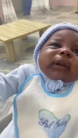 Here's a description of the video:

A close-up video shows a baby wearing a light blue knit hat with a small "Bear" tag. The baby is also wearing a blue-trimmed white bib with a heart design. The baby is crying and appears uncomfortable. A small wooden table is partially visible in the background. The lighting appears to be from indoor sources.