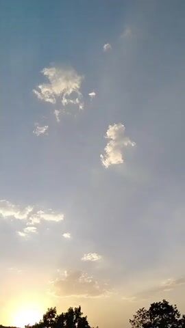 The video displays a sky with scattered clouds, transitioning from a clear blue at the top to a lighter hue near the horizon where the sun is setting. Silhouettes of trees are visible against the sunset glow at the bottom of the frame. The scene is static, with no visible people or objects in motion besides subtle atmospheric changes.