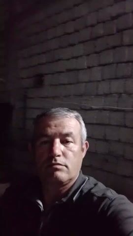 The video features a middle-aged man with short, graying hair and a dark gray shirt. He is standing indoors, and the background appears to be a poorly lit space with brickwork visible. The man raises his right hand and waves in front of him. The lighting is dim. Some wires are also visible above the man. There is music playing in the background.