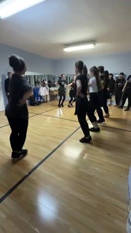 A group of approximately 10 women are practicing a dance in a dance studio. They are all wearing black pants, and most are wearing black shirts. One woman in the middle is wearing a white shirt. They are holding hands and moving in a circle. Another woman stands to the side, observing and demonstrating the dance. There are a few men standing along the wall, watching. The room has a hardwood floor, mirrors along one wall, and a door on another. There are tables with tablecloths and items on them in the background, suggesting a social gathering may follow.